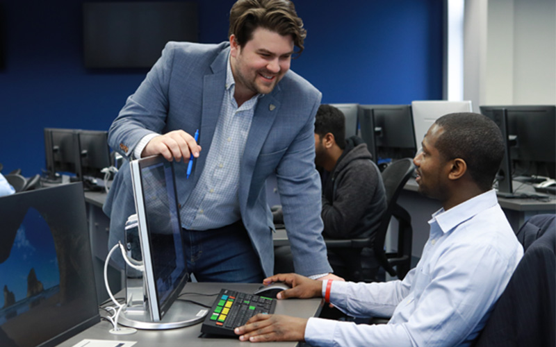 student sitting in front of computer in a computer lab talks to professor