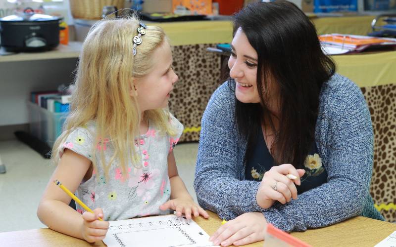 teacher helps a young student sitting at desk on an assignment