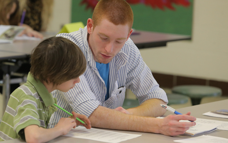 a man sits besides a child at a table in a classroom as the both look over one of the worksheets