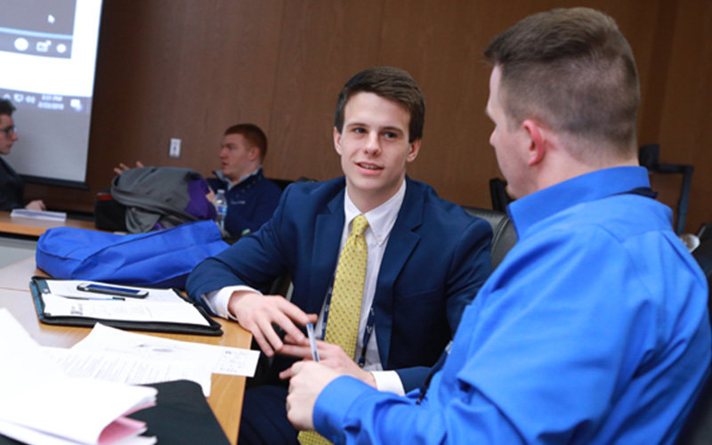 two sales students dressed in suits sit at a table to talk with notes open 