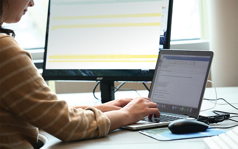 a woman sitting in front of a computer monitor displaying a spreadsheet types onto her laptop