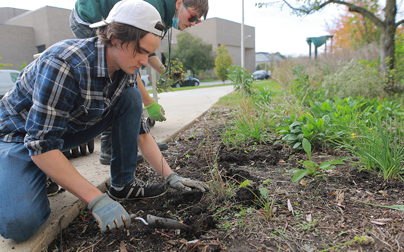 two students outside on the side of the road using gardening tools to dig up dirt and plant different plants
