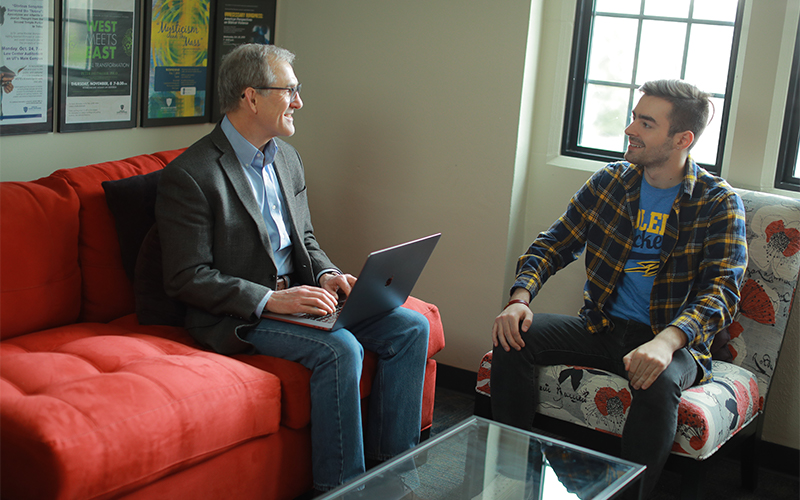 a man sitting on a couch with an open laptop on his lap talks to the man seated adjacently in the chair next to him