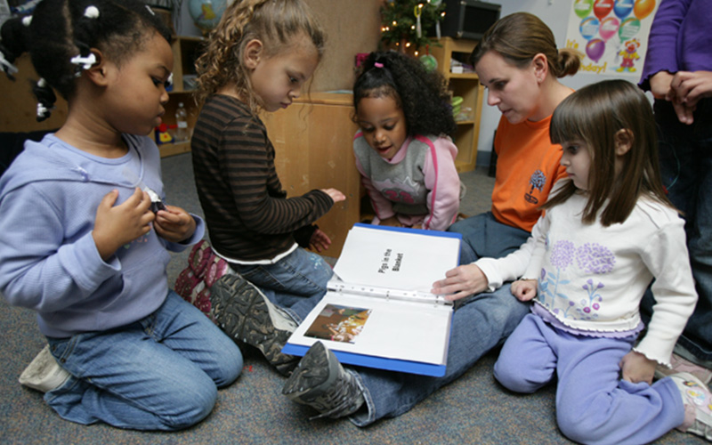 a woman sits on the floor with a small group of young children and reads from a blue binder