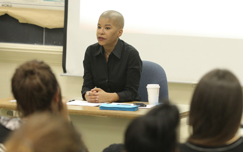 professor sits at desk in front of class while presenting a lecture to the listening audience