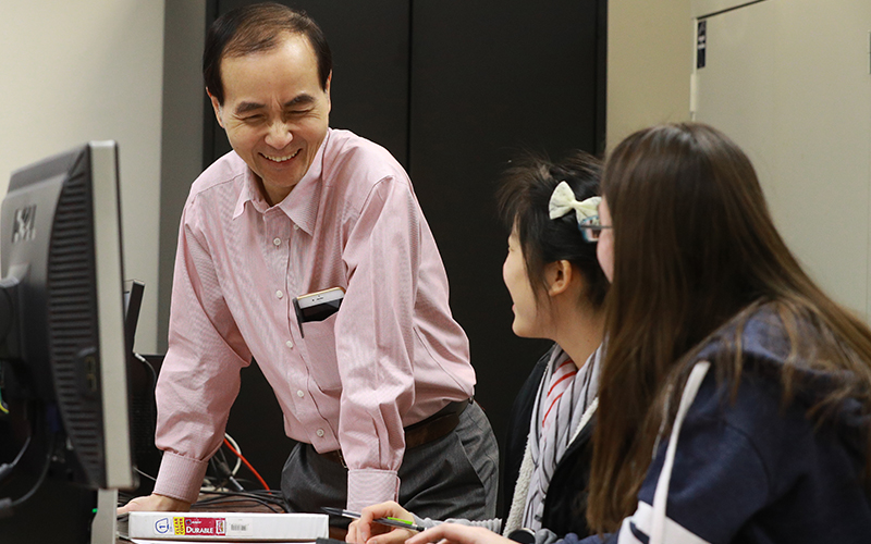 professor smiles and interacts with two students seated at a computer 