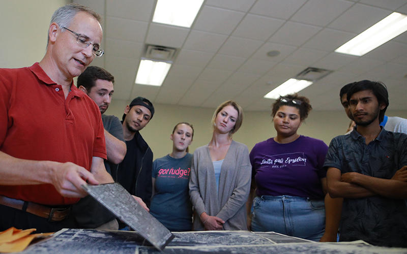 a group of students stand around table and observe as professor holds up a sample