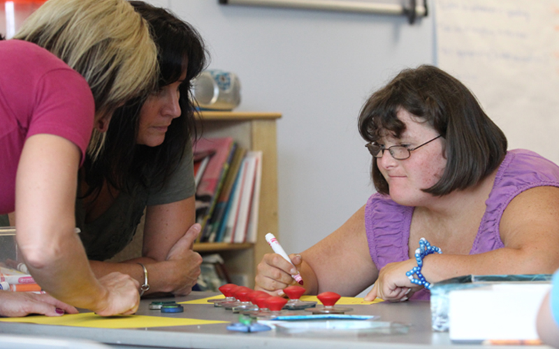 two teachers sit and assist a young student at a table holding a marker