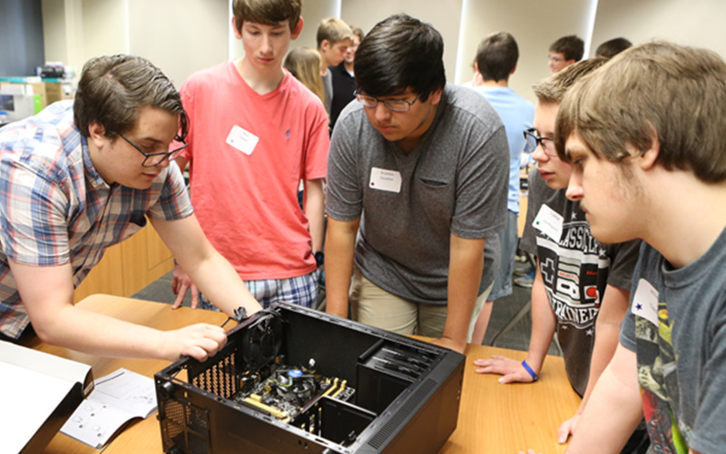 a group of student stand huddled around a table and observe an unassembled computer