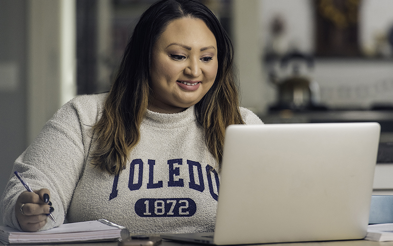 woman wearing a UToledo sweatshirt looks down at her open laptop next to her pen and notebook