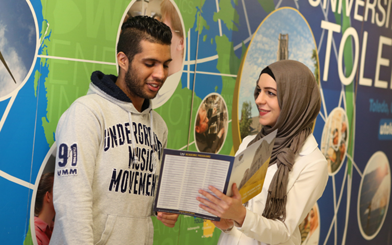 a woman holds up a pamphlet while engaging in converstion in front of a wall featuring a world map 