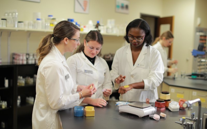 three pharmacy students wearing white lab coats in a pharmacy lab