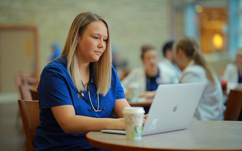 student in blue scrubs sits at table in front of an open laptop