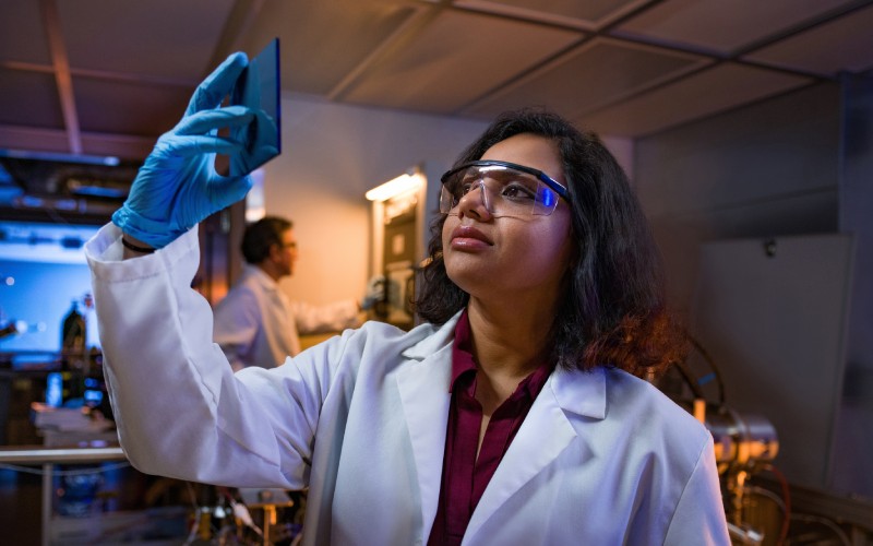 UToledo student in lab coat and protective goggles observing a petri dish