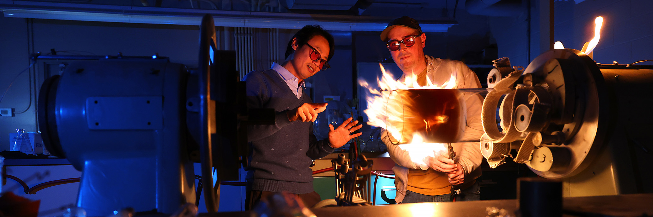 Two people demonstrate a flame-producing machine in a blue-lit laboratory