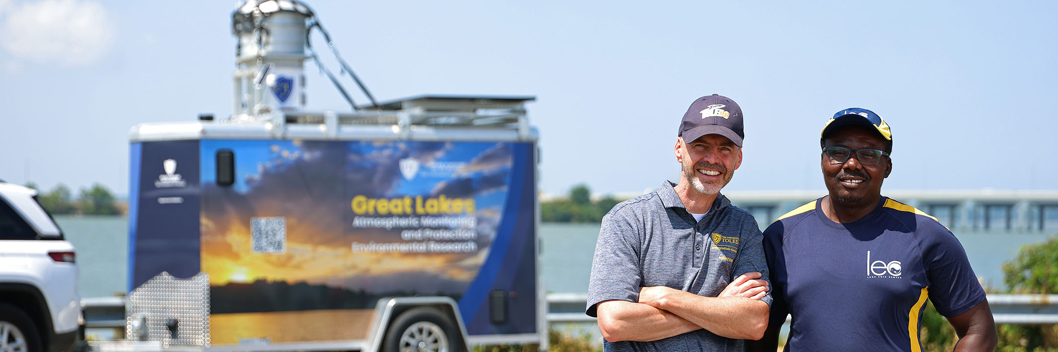 Two men standing in front of a great lakes research trailer in front of a body of water