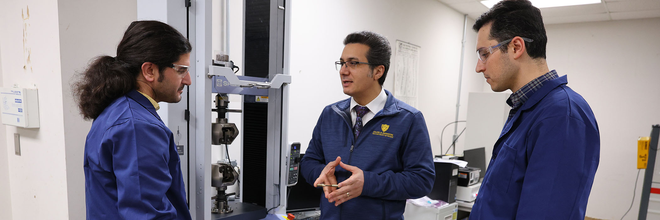 One male researcher explaining something to two others while holding a small piece of equipment in front of a large machine