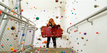 Student standing at the top of a staircase holding open a briefcase as bouncy ball comes tumbling out down the stairs