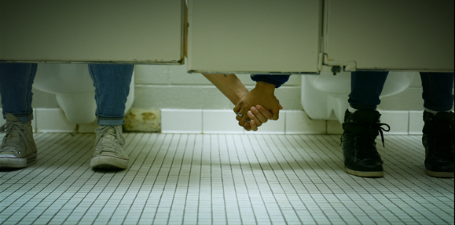 Two people in separate bathroom stalls reaching under the stalls and holding hands in support