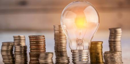 Image of a light bulb sitting upright on a table surrounded by stacks of coins. 455px