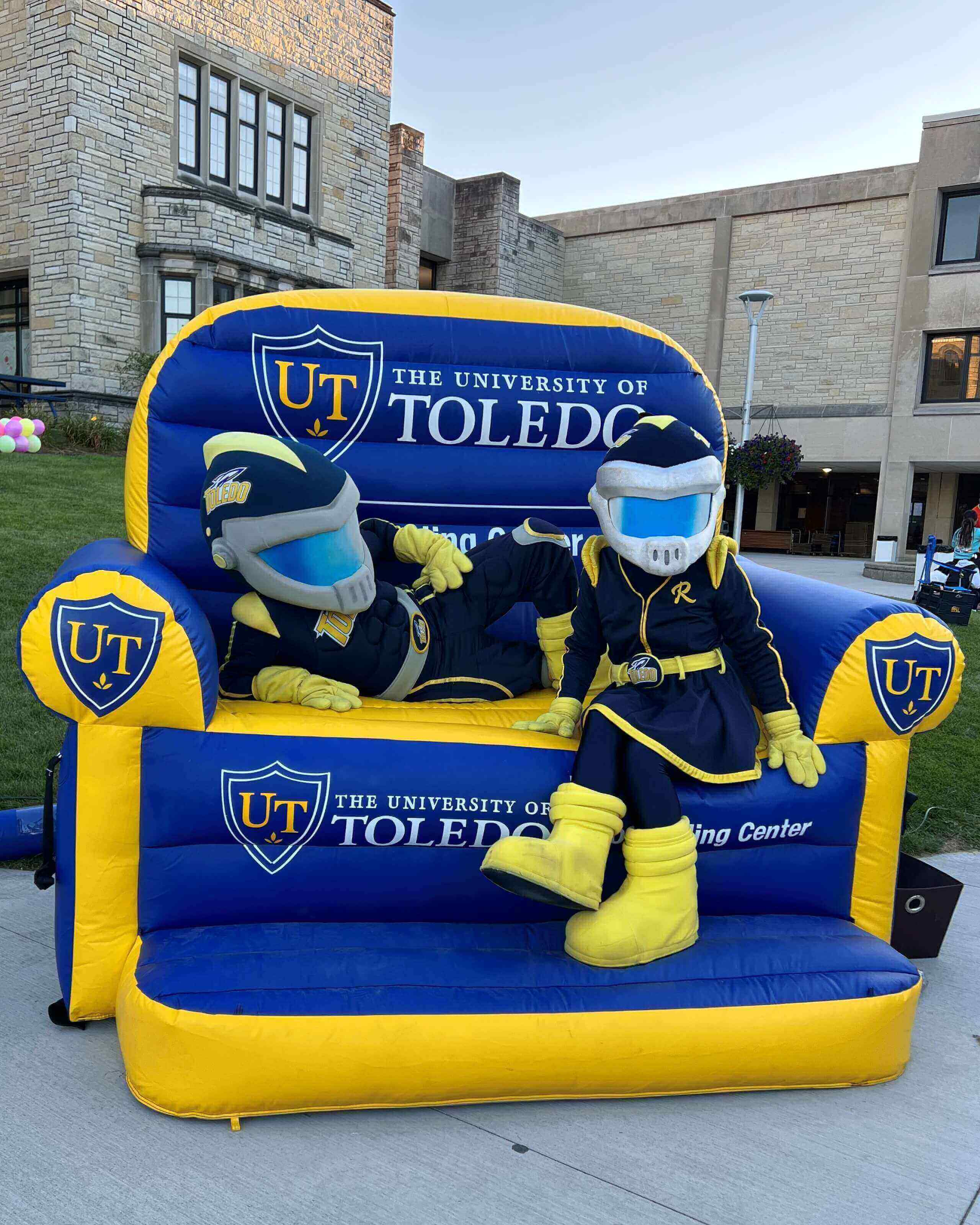Photo of astranaut type mascots, one male and one female, sitting on a large inflatable blue and yellow university of toledo chair. 