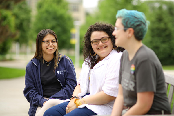Three UToledo students sitting on a bench outdoors smiling and conversing
