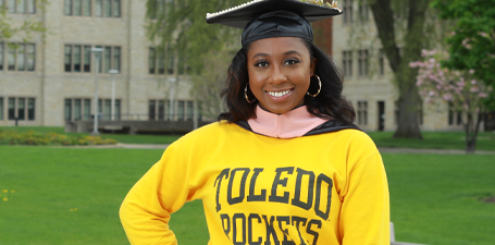 A female, black graduate wearing a graduation cap and Toledo Rockets sweatshirt on campus.