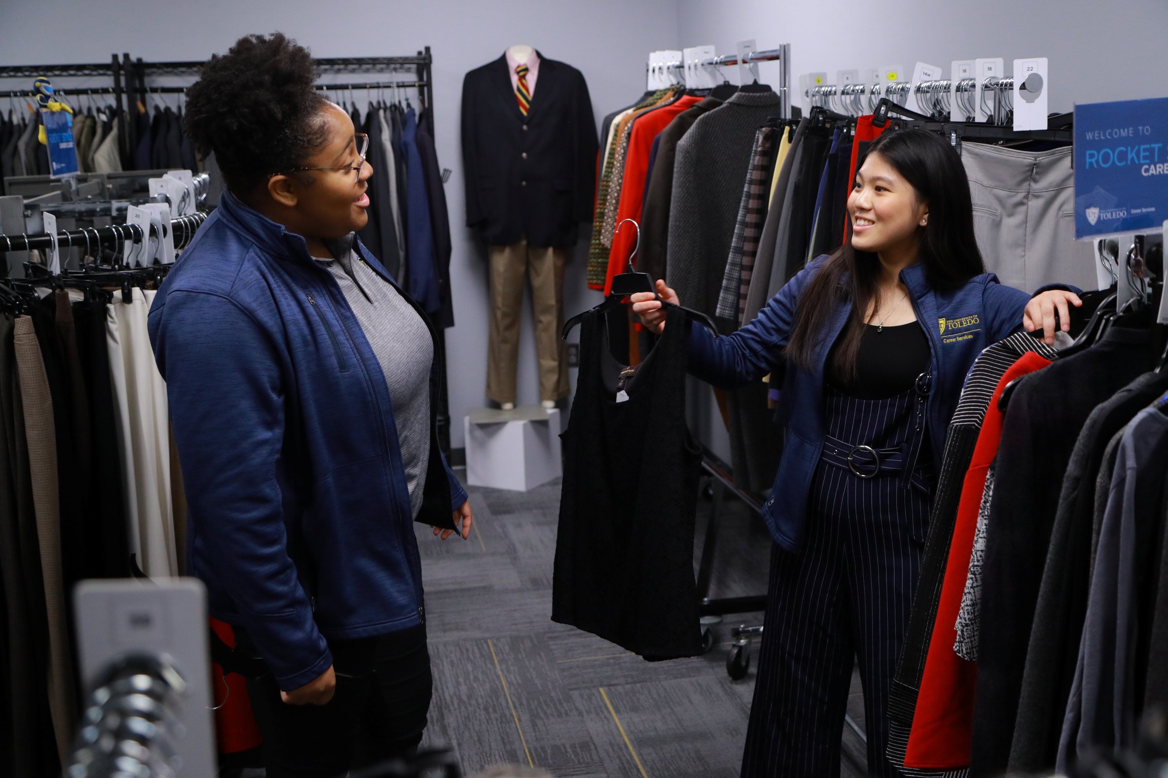 two females standing and talking amongst professional clothing on racks in the Career Closet